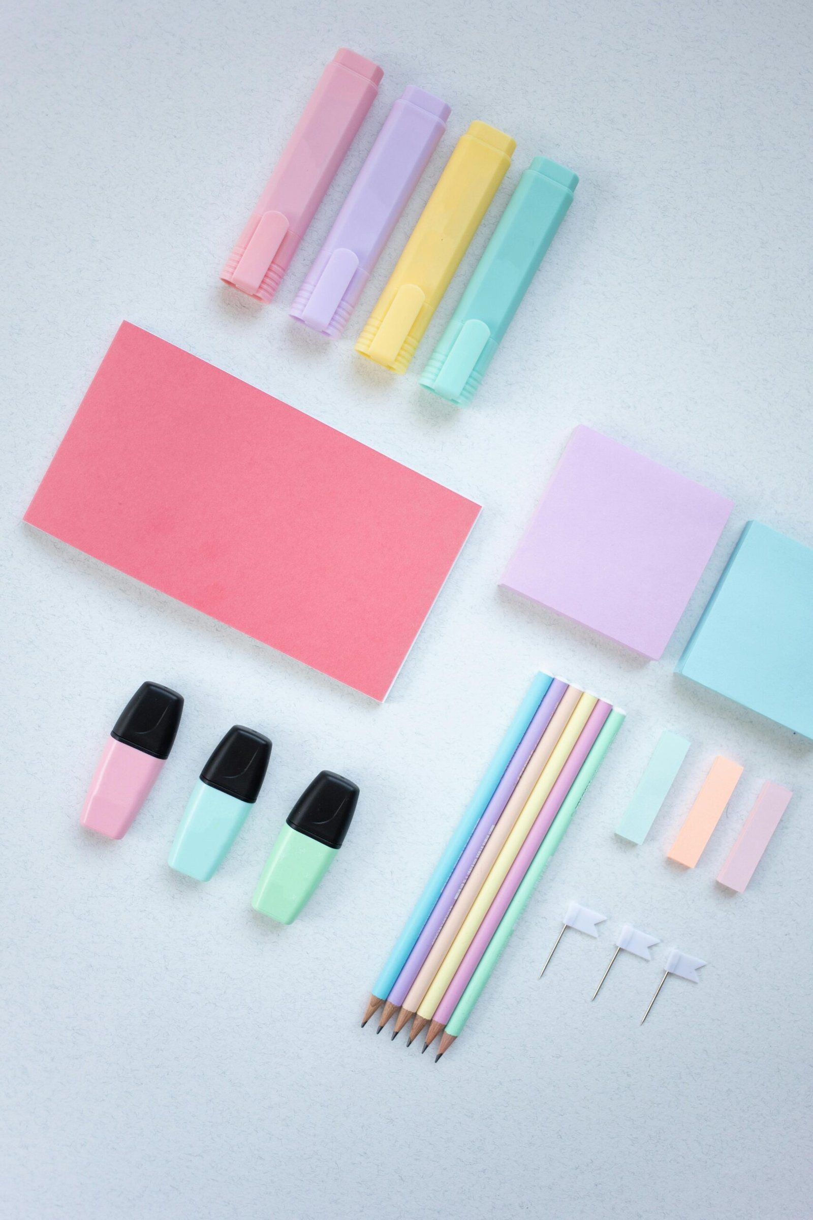 Flat lay of colorful pastel stationery including highlighters, sticky notes, pencils, and push pins on a white surface.